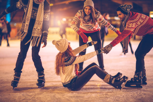 Friends having so much fun while ice skating. Wearing warm clothing. City is decorated with christmas lights.